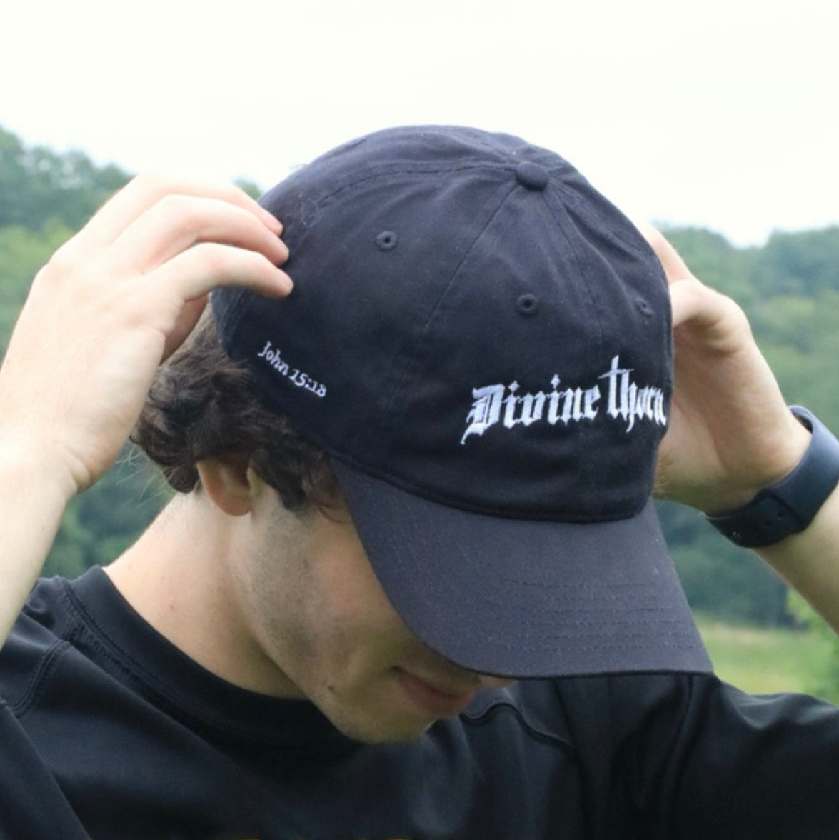 Person wearing a black cap with 'Divine' text and a black shirt with 'West Chester' text, outdoors.