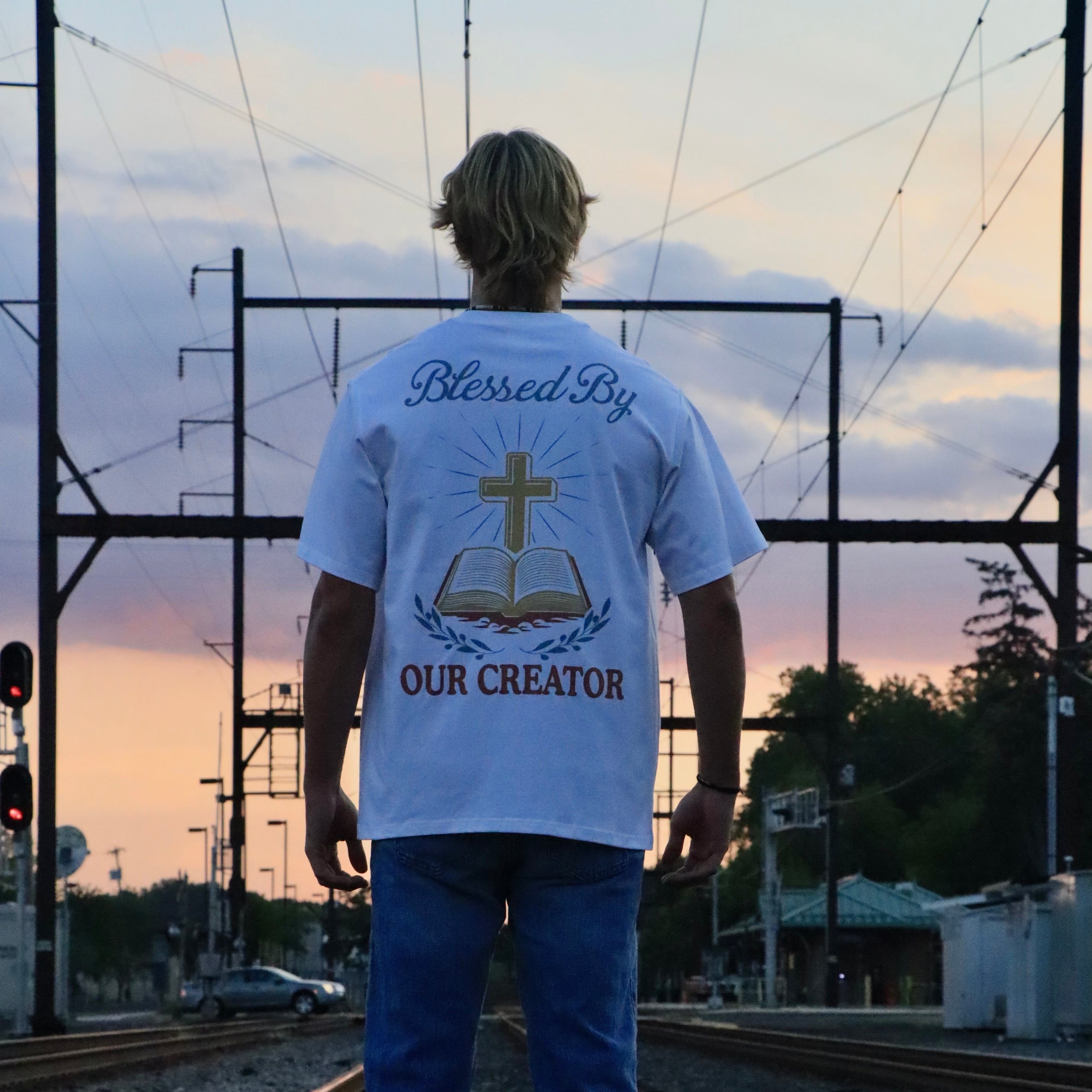 Person wearing a white t-shirt with a religious design and text on a railway track at sunset.