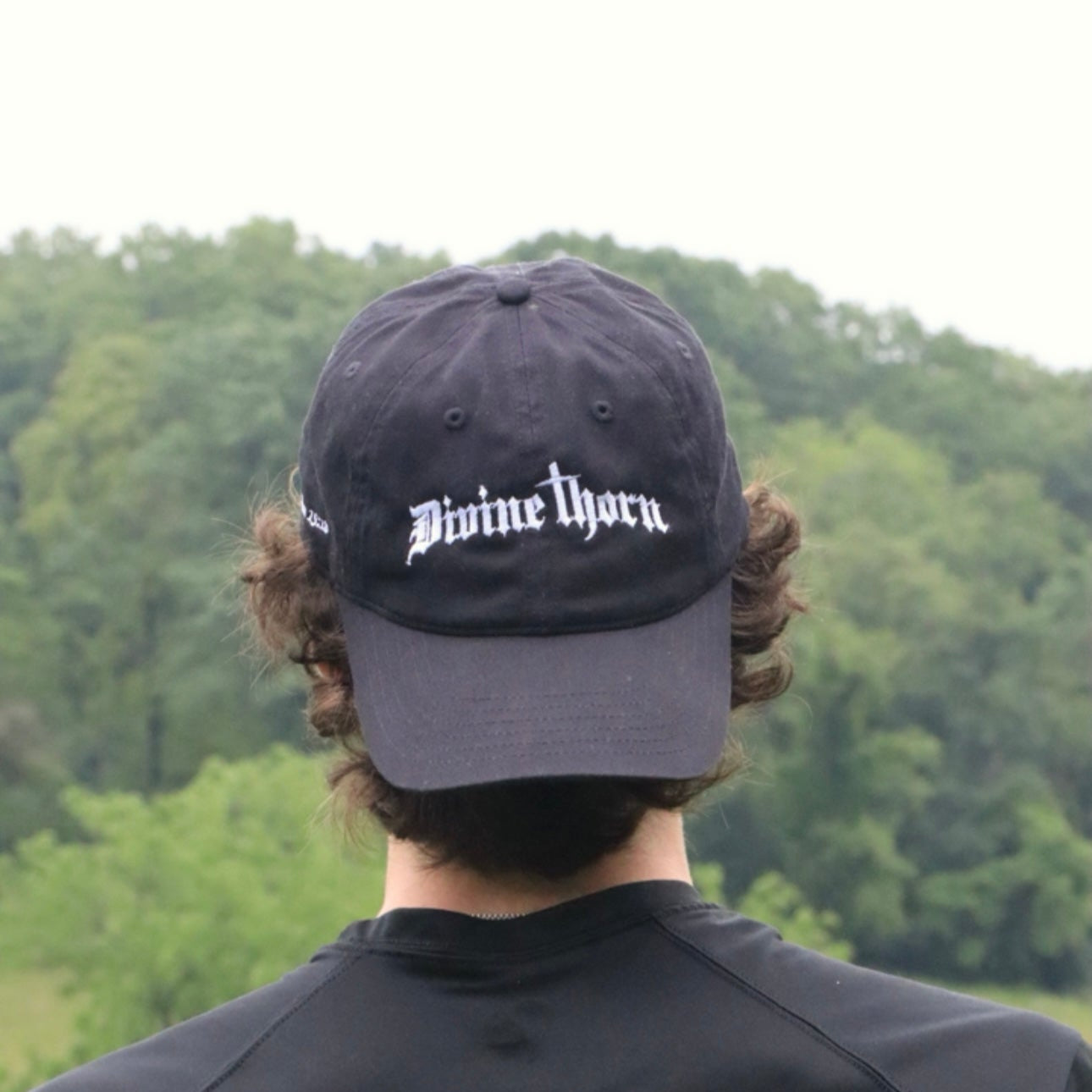 Person wearing a black cap with 'Divine Thorn' logo, standing in front of a forest.