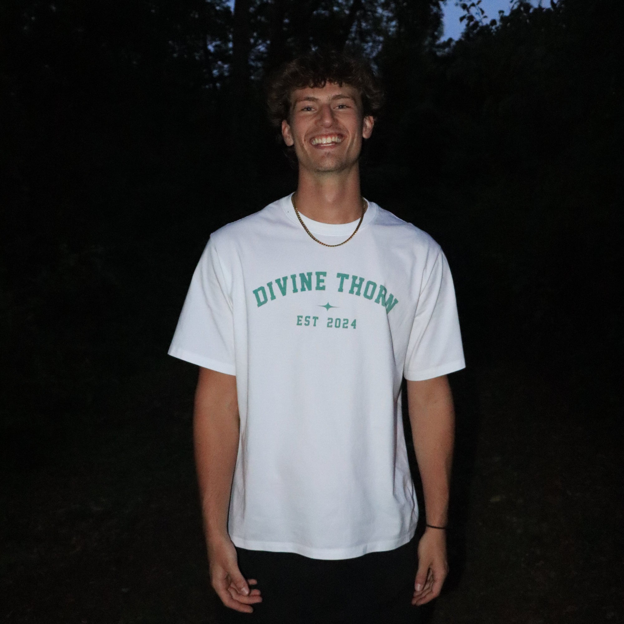 Person wearing a white t-shirt with 'Divine Thorn' text, standing in a dark forest.
