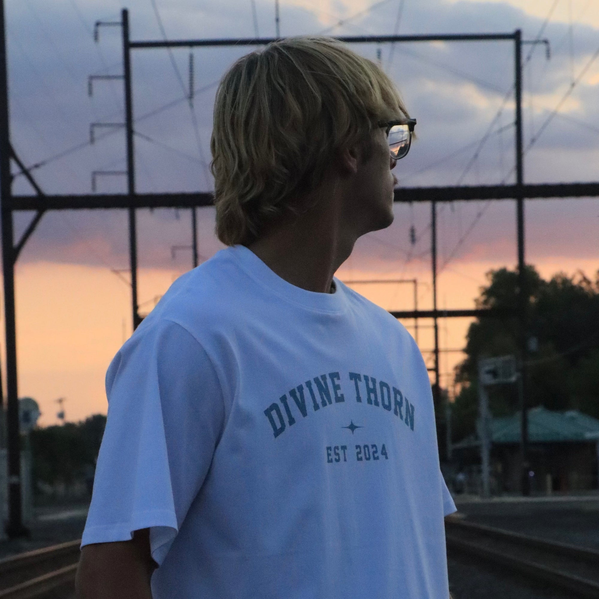 Person wearing a white t-shirt with text standing on train tracks at sunset.